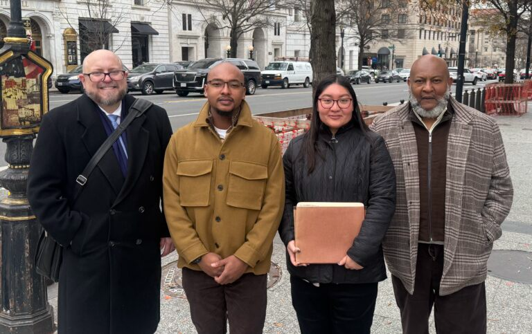 Four adults standing side by side on a city sidewalk, dressed in coats, with buildings, parked cars, and street barriers visible in the background