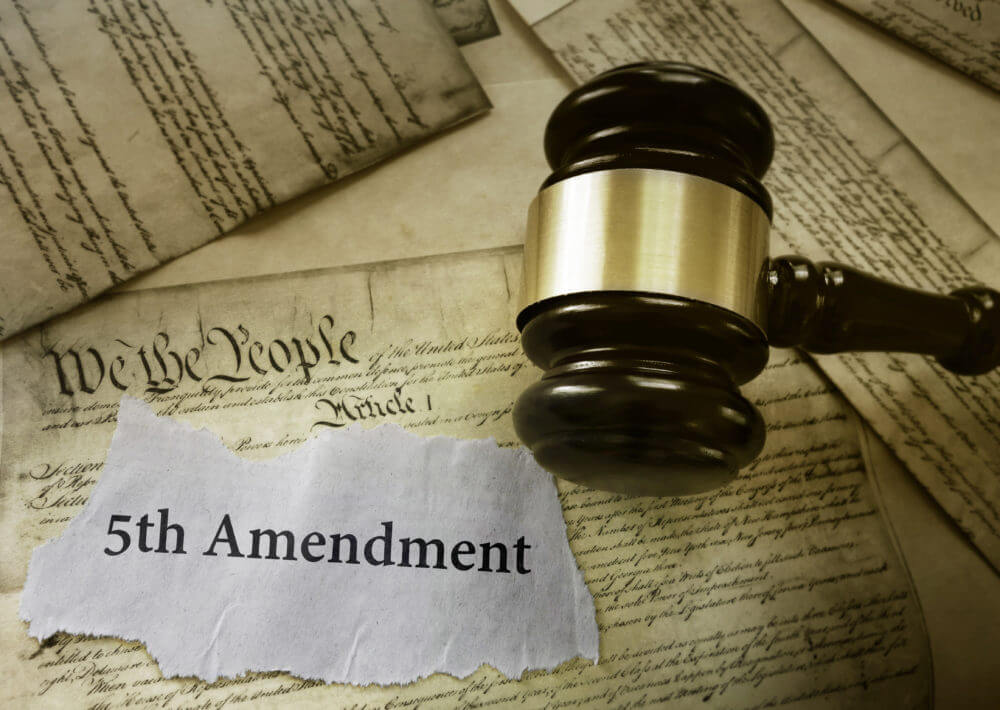 Wooden judge’s gavel resting on aged legal documents with visible historic script, including a torn paper labeled “5th Amendment,” symbolizing constitutional law, legal rights, and courtroom proceedings.