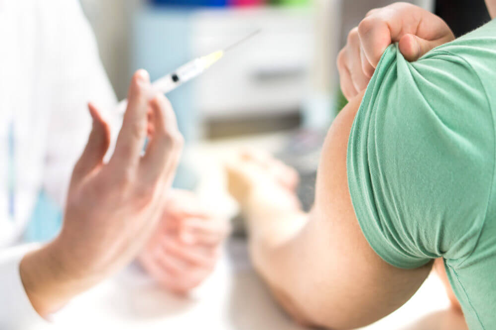 A healthcare professional holding a syringe while preparing to administer an injection into a person’s upper arm, with the patient’s sleeve pulled up and the background softly out of focus.