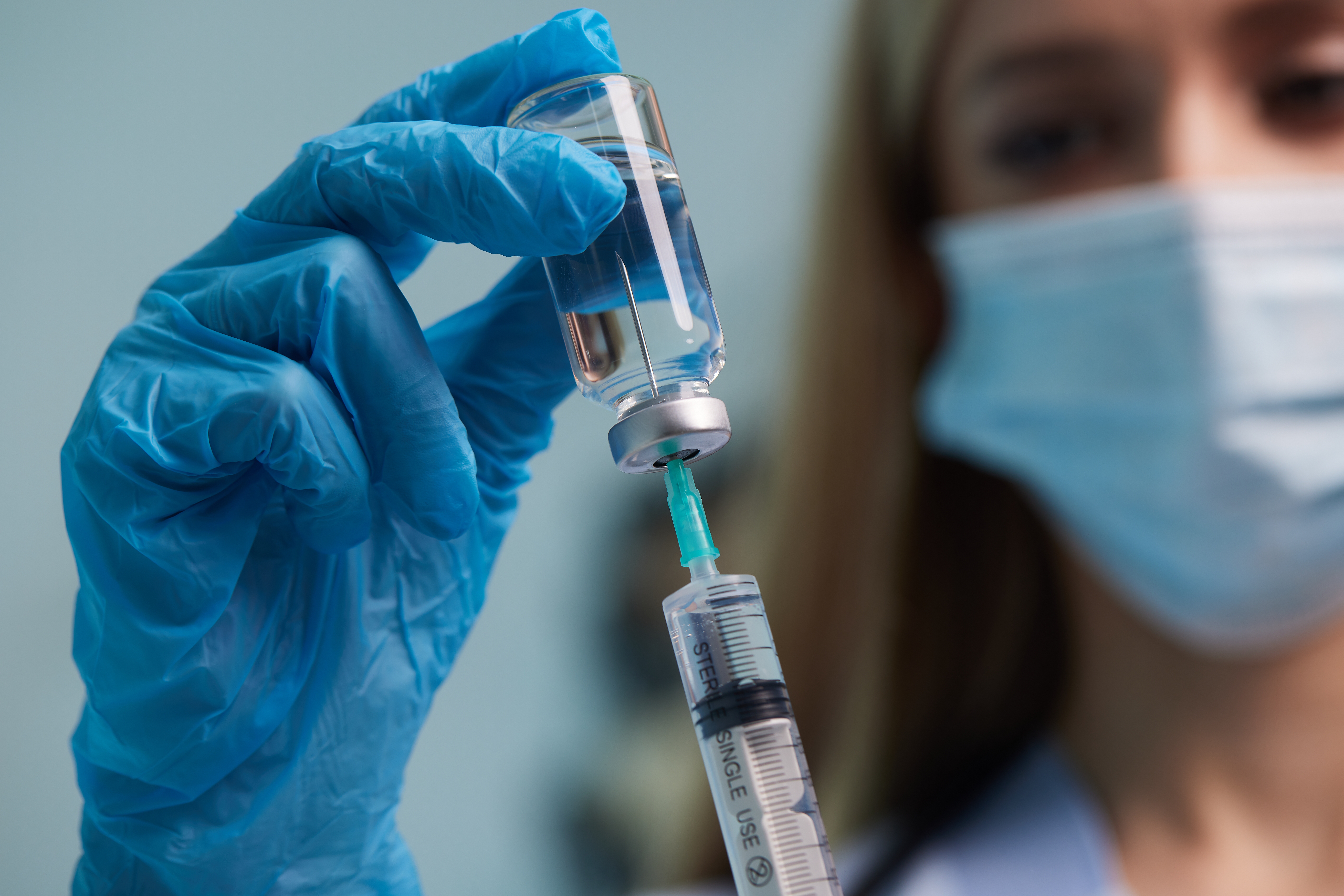 Gloved hand holding a small vial while drawing liquid into a syringe, with a masked medical professional blurred in the background