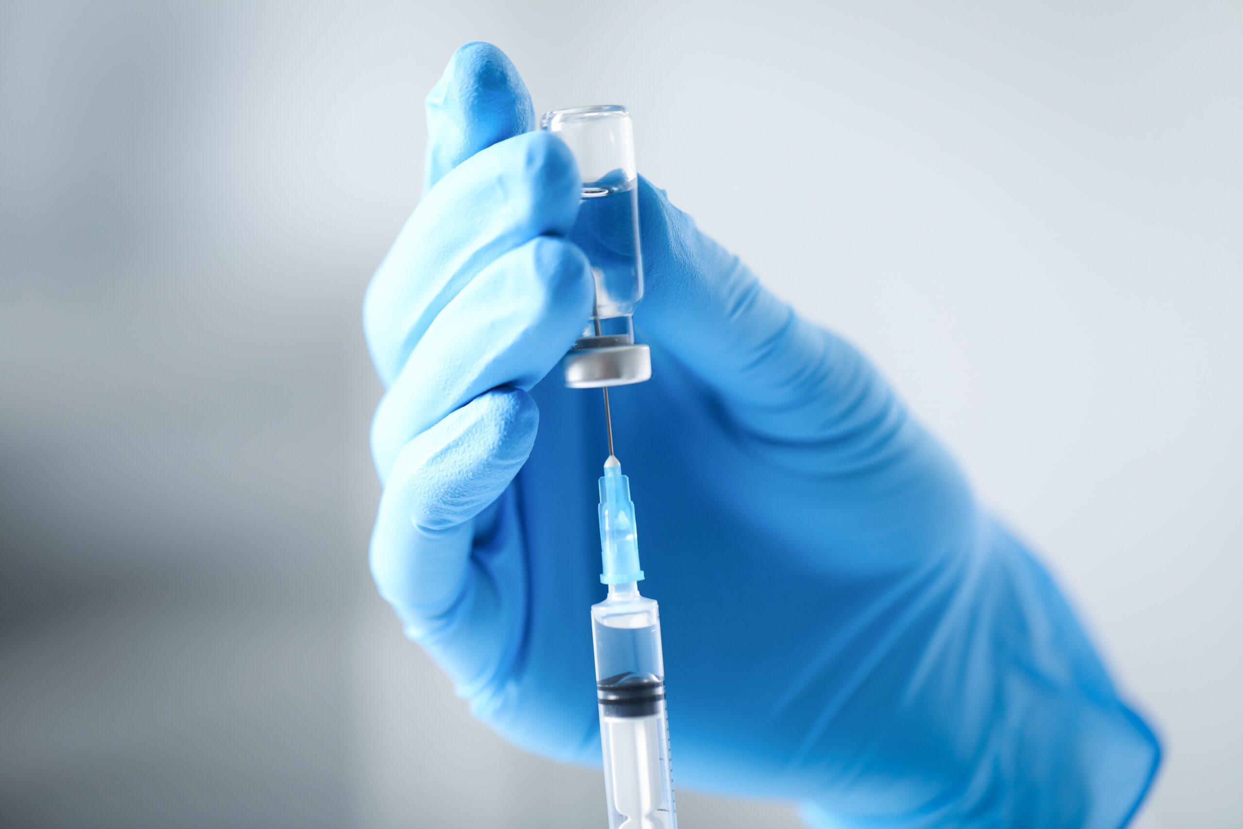 Close-up of a gloved hand holding a vial while a syringe is inserted to draw liquid, set against a clean clinical background